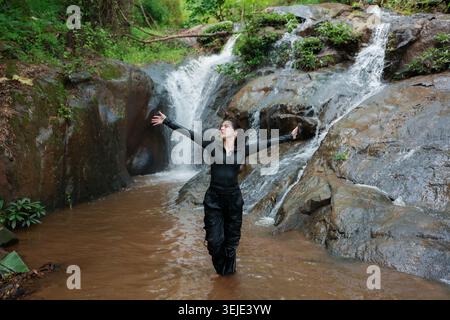 Happy young woman standing in nature waterfall stream forest enjoying outdoor adventure freedom feeling joyful with open arms standing in flowing wate Stock Photo