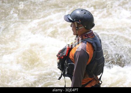 Focused asian man in helmet and life vest ready for rescue or adventure. watchful professional stands with determination beside turbulent whitewater r Stock Photo