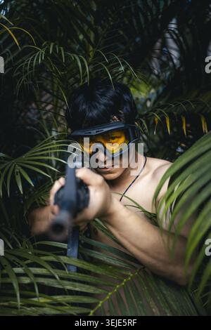 Intense man with gun aiming while hiding in jungle. This soldier uses camouflage with his protective goggle to stalk his target. atmosphere one of sus Stock Photo
