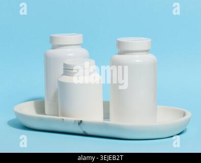 Three white plastic pill bottles of varying sizes stand on a white marble tray against a light blue background Stock Photo