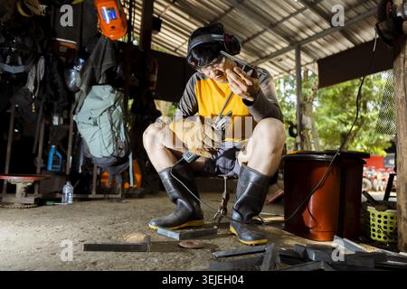 Tired Asian welder and worker taking smoking break from his job. man sits in his workshop, his protective mask up, reflecting on his hard labor and pr Stock Photo