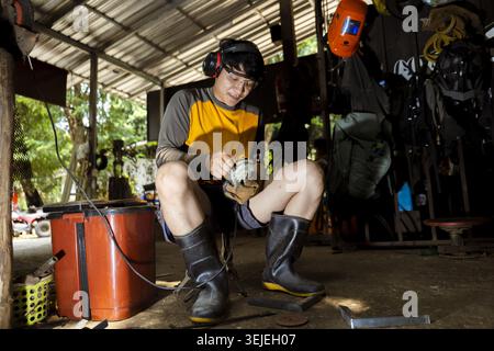 Focused young Asian man in workshop working on manual craft task. He wears earmuffs and boots, showing intense concentration while sitting in his rust Stock Photo