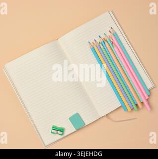 Top view of an open notebook with pastel-colored pencils, green sharpener, and turquoise eraser on a beige background. Stock Photo