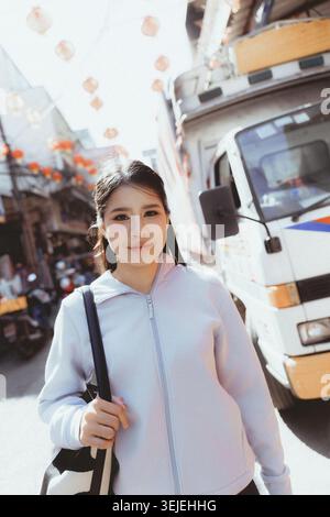 Pleasant young asian woman with long hair enjoys walking on city street. This beautiful person in hoodie looks at camera while exploring urban town ou Stock Photo