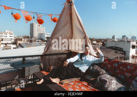 Happy young Asian man relaxing on bohemian rooftop lounge with teepee overlooking city skyline on sunny day. carefree lifestyle and urban travel conce Stock Photo