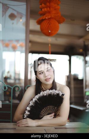Thoughtful Asian woman holding black fan sits in Chinese restaurant. red lantern hangs above, representing tradition and culture during festive celebr Stock Photo