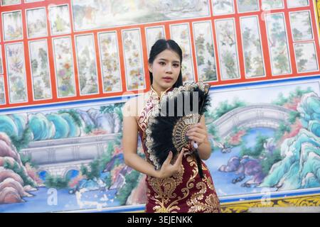 Elegant Asian woman in traditional red qipao dress holding black feather fan poses gracefully. serene young lady inside Chinese temple with mural back Stock Photo