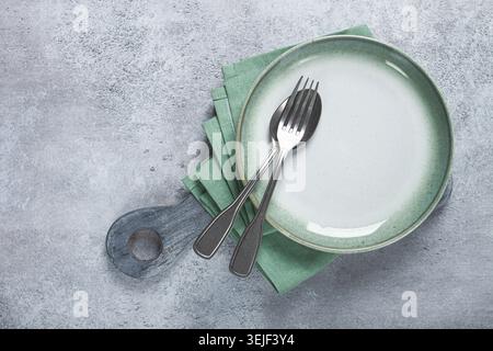 An empty plate and cutlery laid out, on a cutting board, with a green napkin, a gray table, top view, Stock Photo