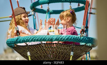 Two girls playing hand clapping game on playground nest swing. Stock Photo