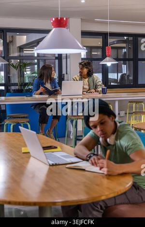 Diverse peers collaborating at co-working space, Hispanic man writing in notebook with laptop Stock Photo