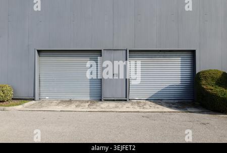 Two metallic roller shutter garage doors on a modern gray facade. Cement sidewalk and asphalt road in front. Background for copy space. Stock Photo