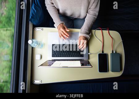 Woman working on laptop while traveling. Computer and smartphone connected to power bank, top view. Stock Photo