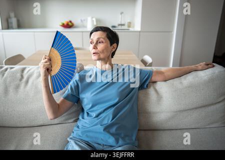 Exhausted mature woman fanning herself at home. Unbearable heat, elderly suffering from stuffy Stock Photo