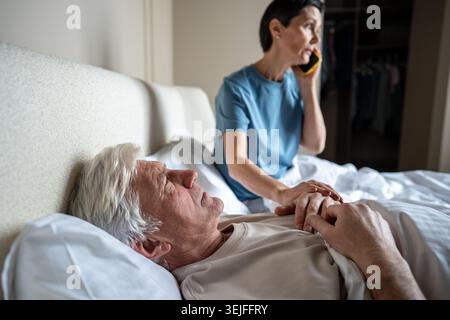 Weak aged man in pain lying in bed, wife dialing ambulance with care. Senior couple domestic sick Stock Photo
