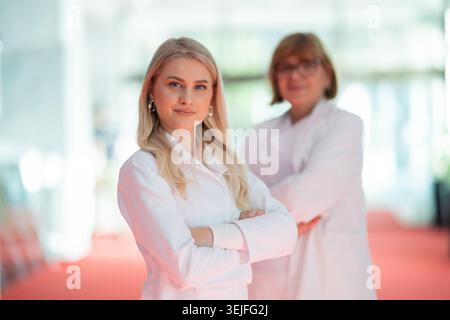 Blonde female doctor in white lab coat standing with arms crossed while senior colleague stands behind her, smiling. Stock Photo