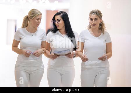 Group of female nurses in white uniforms walk together down a bright hospital corridor, smiling and talking. Stock Photo