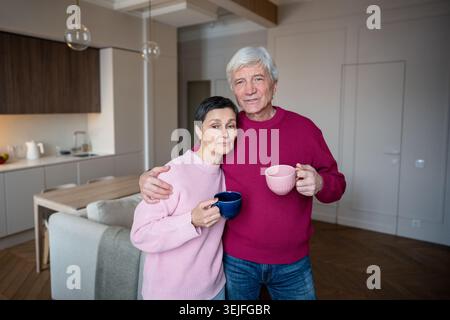 Calm retired wife and husband embracing in morning while drinking tea coffee looking at camera Stock Photo