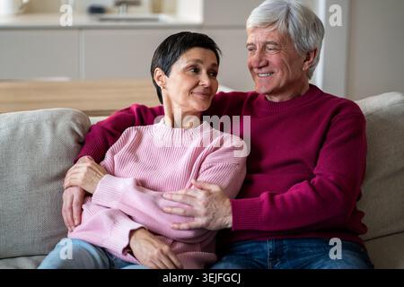 Happy serene elderly couple embracing on sofa at home, wife listen husband with loving attention Stock Photo