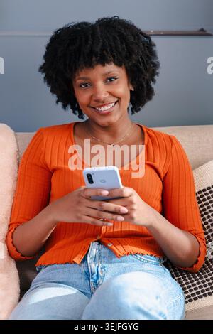 Vertical portrait Cheerful Black woman laughing while using a smartphone on a sofa at home Stock Photo