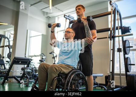 Trainer assists young disabled man in wheelchair on personal session at gym, proper exercises Stock Photo