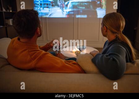 Young couple relaxing at home watching movie on projector while drinking wine on sofa rearview Stock Photo