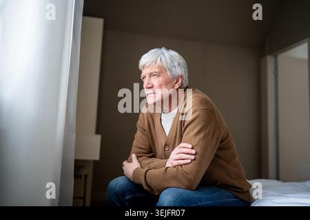 Depressed lonely elderly man look out window sit on bed in nursing home. Suffering unhappy male Stock Photo