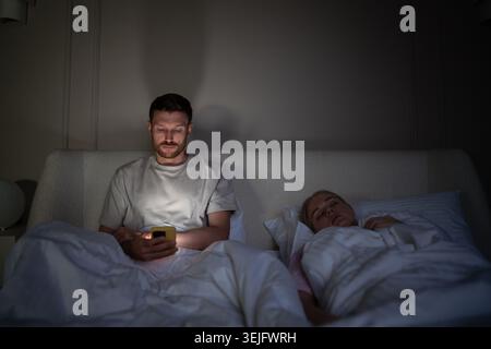 Tired man using smartphone lying in bed at night while wife sleeps beside him in dark bedroom Stock Photo