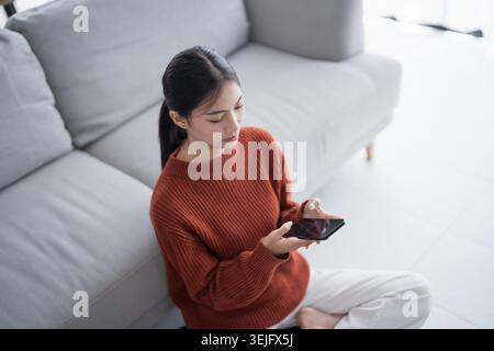 Asian Woman controlling smart home. mobile phone with smart home app in living room. touch screen ai technology. Remote home control system Stock Photo