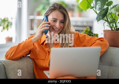 Excited middle-aged woman simultaneously using laptop and talking on phone gossiping with friend Stock Photo