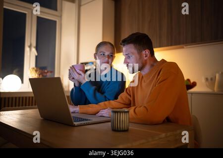 Calm loving couple drinking tea and using laptop together at home in evening, choosing movies Stock Photo