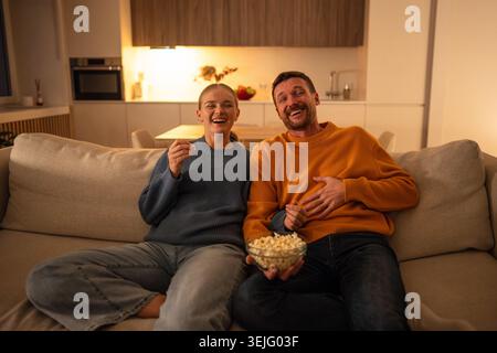 Happy intrigued couple watching movie with popcorn at home cozy together enjoying shared tradition Stock Photo
