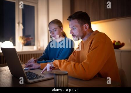Exhausted young couple telecommuting, browsing and working on laptop in kitchen at night. Boyfriend and girlfriend electronic learning together, surfi Stock Photo