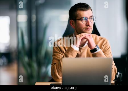 Focused businessman sitting at desk with laptop, thoughtfully analyzing data or planning strategy in a modern office environment, representing concent Stock Photo