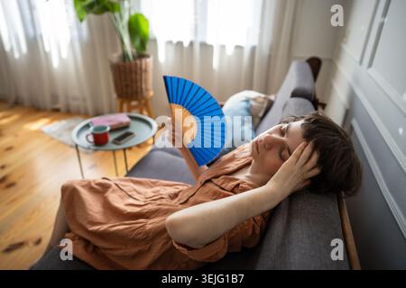 Young woman lying on sofa and waving hand paper fan, suffering from summer heat, feeling headache. Stock Photo