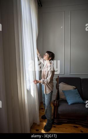 Young relaxed woman standing beside window in cozy living room and opening white curtains in morning Stock Photo
