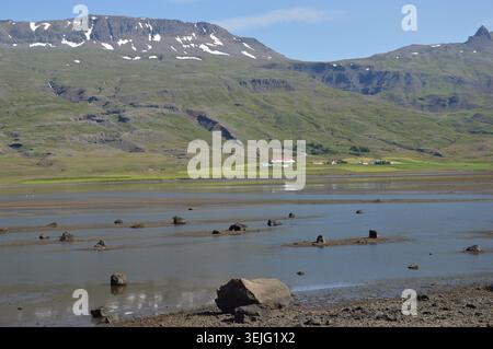 A shallow coastal river estuary with scattered rocks and reflections, featuring a small red-roofed village under snowy mountains. Stock Photo