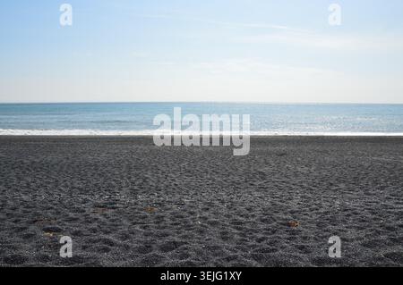 A minimalist landscape view of a dark volcanic pebble beach meeting a calm blue ocean under a bright sky. Stock Photo
