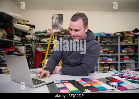 Fashion designer using laptop with fabric swatches in workshop, textile business owner working at his desk, checking inventory, Stock Photo