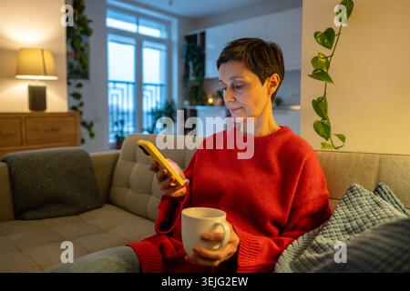 Senior woman resting on couch in living room, holding coffee mug and scrolling media on smartphone. Stock Photo