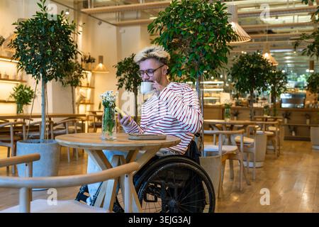 Thoughtful dreamy disabled man in wheelchair drinking coffee at cafe table, using mobile phone Stock Photo