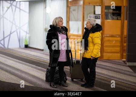 Two older women dressed in winter clothing standing in a hotel lobby and having a friendly conversation next to their luggage. Concept of travel, frie Stock Photo