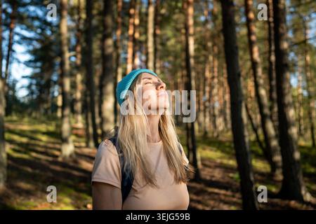 Relaxed woman resting with closed eyes warming in sunlight during forest walk. Meditating in nature Stock Photo