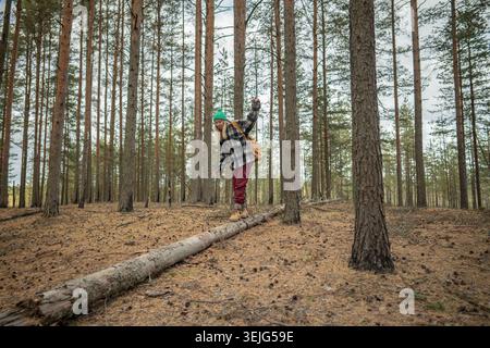 Female backpacker balancing on fallen tree trunk with backpack during autumn hike in pine forest Stock Photo