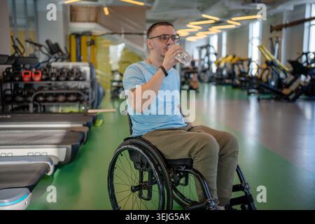 Disabled sportsman in wheelchair resting during powerful workout in gym, drinking water from bottle. Stock Photo