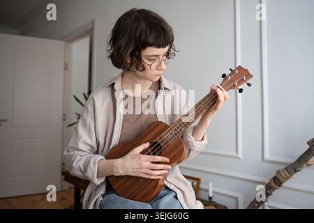 Focused young woman in glasses learning ukulele chords at home, intent on musical progress Stock Photo