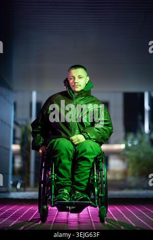 Confident calm young man with disability in wheelchair in color rgb neon light posing at camera. Stock Photo