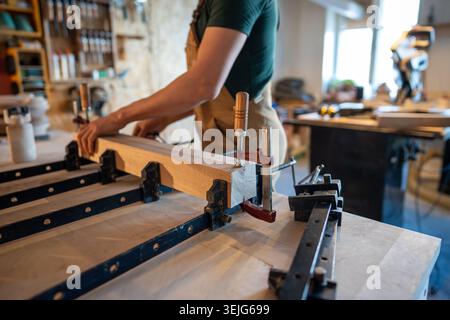 Man carpenter working in carpentry workshop, compressing glued wooden blocks at table with clamps. Stock Photo