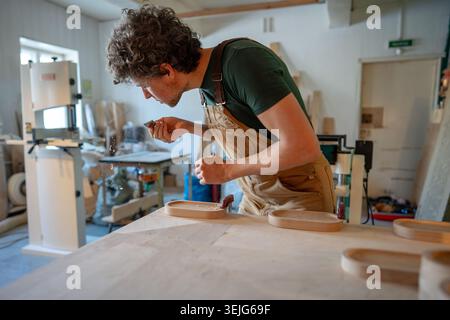 Man carpenter blows dust off chisel blade, carefully sanding coasters for glasses with sand paper. Stock Photo