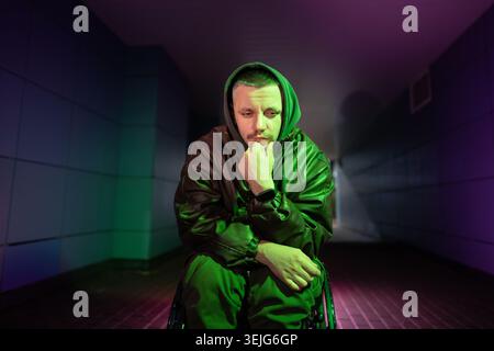 Sad disabled man in wheelchair thinking about personal crisis, sits alone in rgb neon color light. Stock Photo