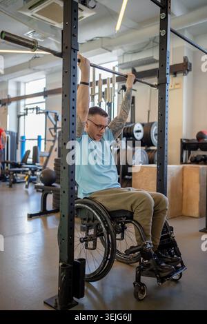 Man athlete in wheelchair doing vertical pull ups exercise using adaptive sports equipment at gym. Stock Photo
